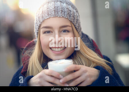 Portrait von junge, blonde Frau in Hut und Strickjacke halten Kaffee, lächelnd und Blick in die Kamera, über Outdoor-unscharfen Hintergrund hautnah Stockfoto