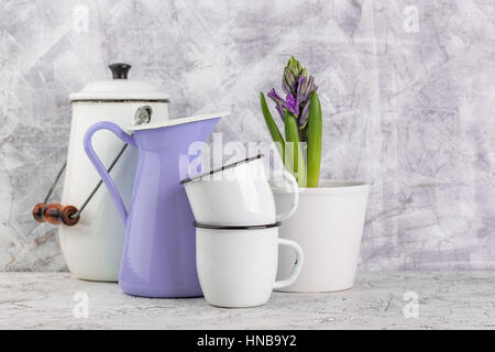 Zwei weiße Emaille Becher, violettes Glas, weiß Can und blühenden Blume Hyazinthe im Topf auf hellem Hintergrund Stockfoto