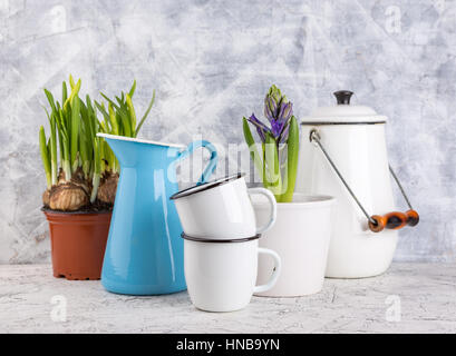 Zwei weiße Emaille Becher, blaues Glas, weiße Dose und Frühling Blumen im Topf auf hellem Hintergrund Stockfoto