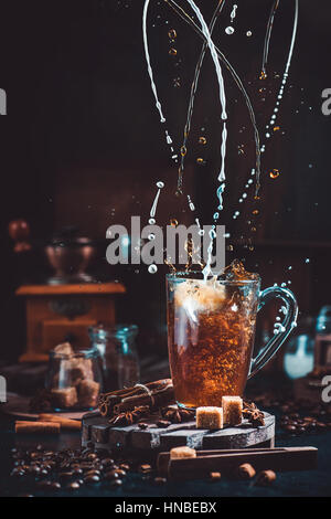 Verdrehen, Milch und Kaffee in einer transparenten Tasse auf einem dunklen Hintergrund mit Schleifer, Löffel, Zimt und Kaffee Kaffeebohnen spritzt Stockfoto