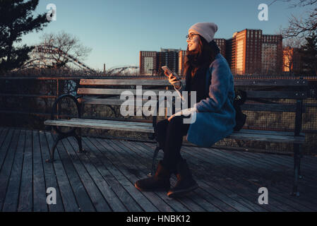 Junge attraktive Mädchen in warme Kleidung halten Handys beim Sitzen auf Bank und wegsehen Stockfoto