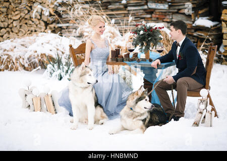 Braut und Bräutigam im Winter mit zwei Hunden Malamute am Tisch sitzen. Auf dem Tisch liegen Wein Kelche, Kerzen und Bouquet. Stockfoto