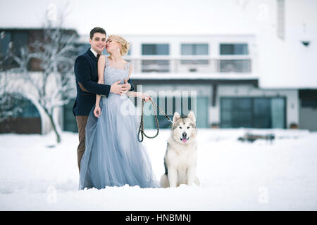 Braut und Bräutigam im Winter mit Hund Malamute auf Schnee und Zuhause Hintergrund. Bräutigam umarmt Braut, und sie sieht ihn zärtlich. Stockfoto