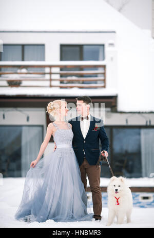 Braut und Bräutigam im Winter mit Hund auf Schnee und Zuhause Hintergrund. Bräutigam umarmt Braut und sie schauen zärtlich gegeneinander. Stockfoto