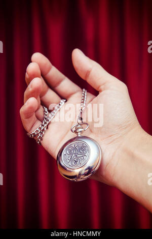 Männliche Hand, die eine Vintage Silber Taschenuhr mit Gravur auf dem Deckel, roten unscharfen drapieren Hintergrund, ausgewählte Fokus geschlossen Stockfoto