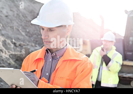 Ingenieur in Zwischenablage auf Baustelle mit Kollegen im Hintergrund schreiben Stockfoto