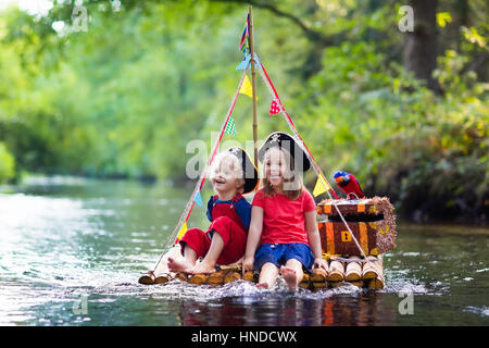 Kinder gekleidet in Piraten Kostüme und Hüte mit Schatztruhe, Ferngläser und Schwerter spielen auf hölzerne Floß Segeln in einem Fluss an heißen Sommertag. Pira Stockfoto