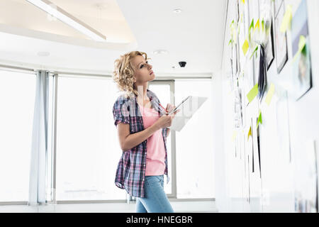 Kreative Geschäftsfrau Blick auf Papiere fest an Wand beim Schreiben von Notizen im Büro Stockfoto