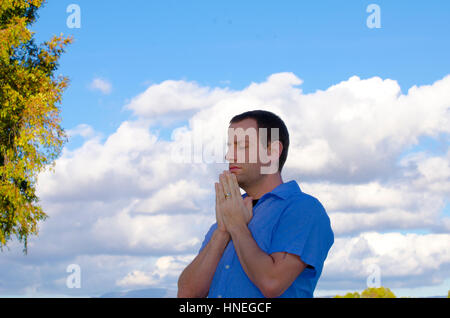 Mann in einem blauen Hemd draußen und betete mit den Wolken und Bäumen im Hintergrund. Stockfoto