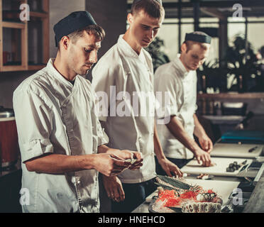 Drei junge Köche in weißer Uniform gekleidet dekorieren fertig Gericht im Restaurant. Sie arbeiten an Maki-Rollen. Traditionelles japanisches Sushi Zubereitung eingestellt Stockfoto