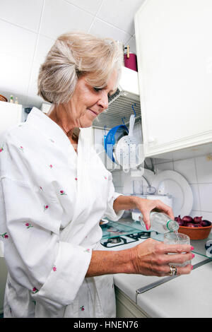 Ältere Frau gießt Wasser in Glas im stehen am Küchentisch Stockfoto