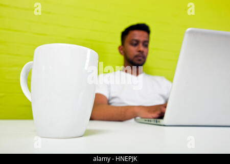 Indischen Mann arbeitet an seinem Schreibtisch mit Becher im Fokus im Vordergrund Stockfoto