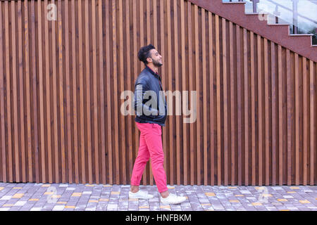 Hübscher junger Erwachsenen muslimischen Mann, Geschäftsmann Spaziergänge langsam entlang der Treppe, sieht sich um und lächelt, hält Hände in den Taschen Jacken, wirbt abwaschen Stockfoto
