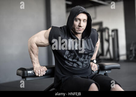 Brutaler Mann mit starken Muskeln tut auf der Bank in der Turnhalle heben. Er trägt schwarze Kapuze ärmellos und schwarzen Shorts. Mann schaut in die Kamera. Hori Stockfoto