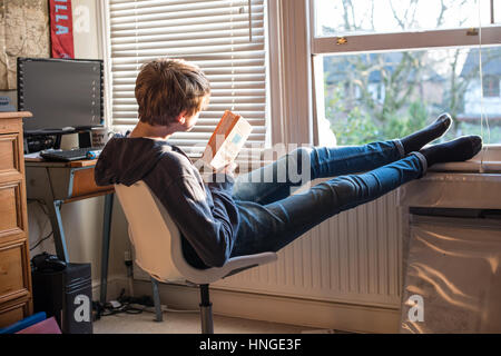 Weiße Teenager in Jeans und ein Schweiß-Top durch seine offene Schlafzimmerfenster mit seinen Füßen sitzend von seinem Schreibtisch lesen und Revision für GCSE Prüfungen