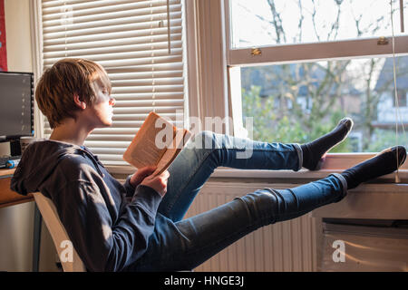 Weiße Teenager in Jeans und ein Schweiß-Top durch seine offene Schlafzimmerfenster mit seinen Füßen sitzend von seinem Schreibtisch lesen und Revision für GCSE Prüfungen Stockfoto