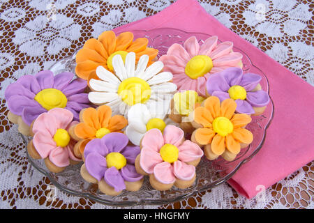 Ausgefallene hausgemachte Frühlingsblumen Zucker Cookies mit königlicher Zuckerglasur Blütenblätter auf geleitet. Serviert auf einem Glas Platte weißer Spitze Tuch rosa Serviette Stockfoto