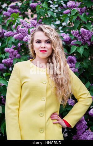 Blonde Mädchen in einem gelben Mantel steht vor dem Hintergrund der blühenden Flieder im Frühjahr Stockfoto