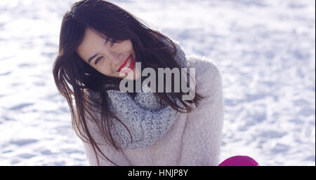 Lächelnde junge Frau in Pullover saß auf Schnee schiefen Kopf zur Seite Stockfoto