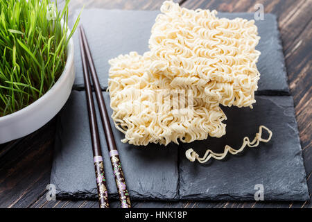 Trocken Sie Ramen-Nudeln auf einem schwarzen Steinplatten mit dekorativen Stäbchen und grünen Kräutern in einer Schüssel Stockfoto