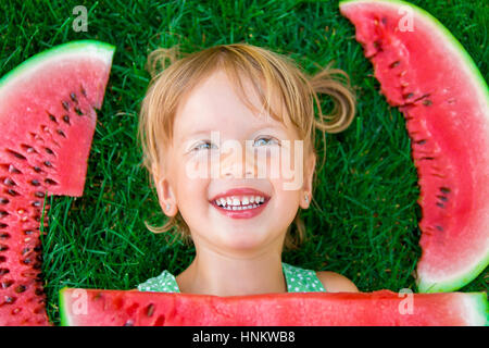 Glücklich kleine blonde Mädchen mit großen Scheibe Wassermelone im Sommer auf dem Rasen liegen. Lächeln auf den Lippen. Stockfoto