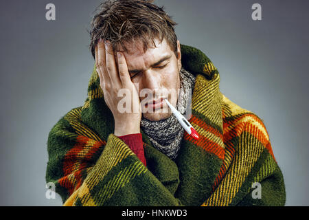 Junge Mann leidet unter Kopfschmerzen. Foto von kranker Mann in Decke gehüllt mit einem Thermometer im Mund. Stockfoto