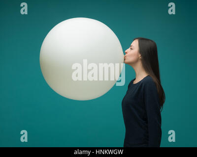 lustige junge Frau mit langen dunklen Haaren aufblasen riesige Ballons auf blauem Hintergrund Stockfoto