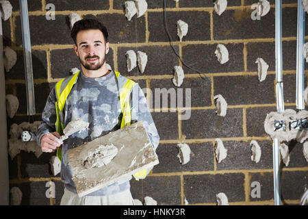 Ein Gipser, ein Bauherr mit Gipskartonplatten und Verbreitung Kelle vor einer Wand Betonklotz. Stockfoto