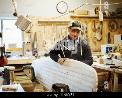 Mann stehend in einem Workshop Smoothng und Schleifen eine hölzerne Surfbrett. Stockfoto