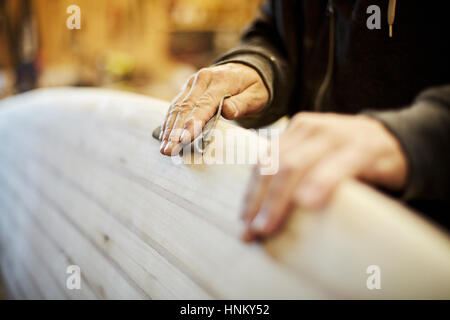 Mann in einer Werkstatt Schleifen den Rand eines hölzernen Surfbretts. Stockfoto