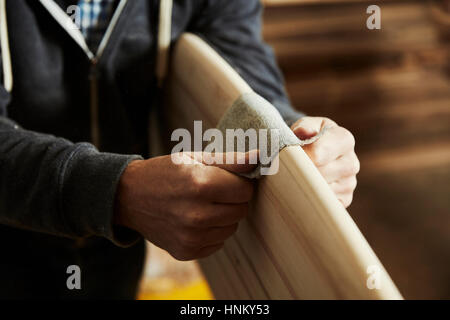 Mann in einer Werkstatt Schleifen die abgerundete Kante eines hölzernen Surfbretts. Stockfoto