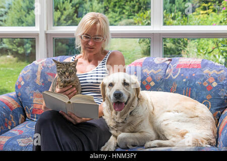 Blonde mittlere gealterte Frau sitzt auf einer Couch und ein Buch zu lesen. Gestreifte Katze und Golden Retriever Hund sitzen mit ihr zusammen. Die Haustiere sind loo Stockfoto