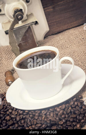 Schrägen Blick auf das Vintage Foto Tasse Kaffee stehen auf dem Leinensack. Es sind Kaffeebohnen verstreut. Der Teil des manuellen Kaffee gri Stockfoto