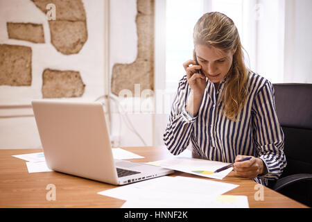 Junge Geschäftsfrau, die Diskussion über ein Diagramm mit jemandem am Telefon während darauf mit einem Stift in ihrer linken Hand zeigen Stockfoto