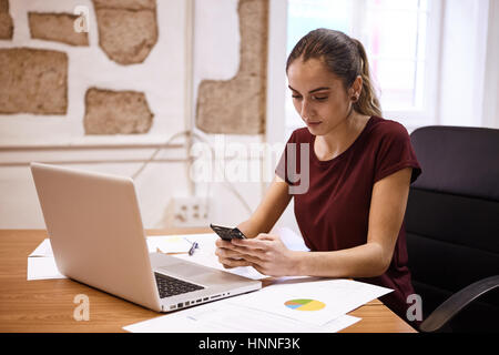 Hübsche junge geschäftsfrau auf Ihr Handy suchen mit ihrem Laptop öffnen vor ihr und einige Kreisdiagramm auf Ihrem Schreibtisch Stockfoto