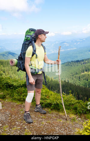 Junge Wanderer mit Bandage am Knie und Spazierstock in der Hand in Bergen. Männer in kurzen Hosen auf Berg mit umschlossenen verletzten Knie mit weißen Sportboykott Stockfoto