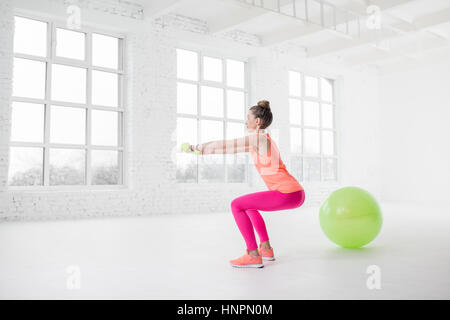 Junge Frau in bunte Sportbekleidung mit Hanteln in der weißen Turnhalle mit Fitness-Ball auf dem Boden hocken Stockfoto
