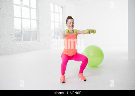 Junge Frau in bunte Sportbekleidung mit Sonnenschirmständer in der weißen Turnhalle mit Fitness-Ball auf dem Boden hocken Stockfoto