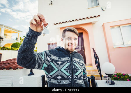 Glücklicher lustiger Mann hält Hausschlüssel auf Haus geformt Schlüsselbund vor ein neues Zuhause Stockfoto