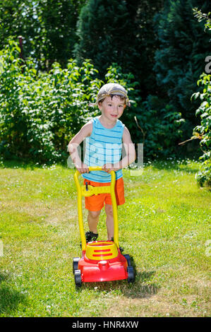 Fröhlicher kleiner Junge Hilfe bei der Gartenarbeit mit seinem Rasenmäher auf sonnigen Tag mit abisolierten T-shirt und Taschentuch mit kleinen Knotts auf Kopf Stockfoto