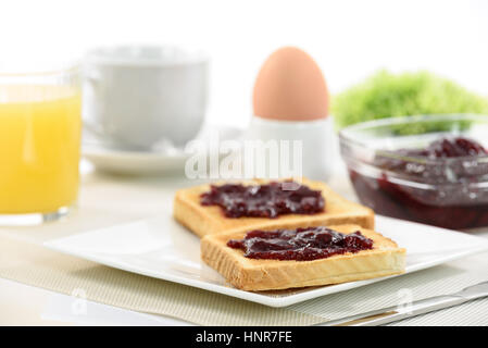 Knuspriger Toast mit Himbeermarmelade auf einen Teller und ein weich gekochtes Ei neben Orangensaft und eine Tasse Kaffee und Äpfel auf weißem Hintergrund. Stockfoto