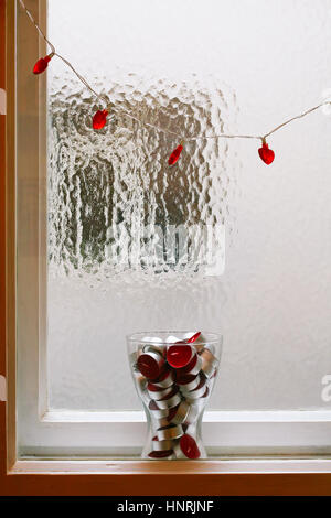 Rotes Herz geformt, Lichterketten und Glasvase voller roter Teelichter zwischen Fensterrahmen. Stockfoto