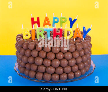 Whopper einer Schokolade Kuchen mit Süßigkeiten Malz Kugeln und Happy Birthday Kerzen dekoriert. Blaue Fläche gelben Hintergrund. Schnelle einfache Hausgemachte Stockfoto