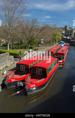 Landschaftlich sonnige Aussicht auf Person, die Hund auf dem Pfad und leuchtend roten Kanal Mietboote vor Anker - Leeds-Liverpool Canal, Skipton, Yorkshire, England, Großbritannien. Stockfoto