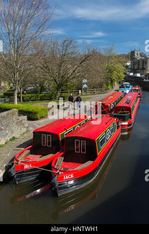 Landschaftlich sonnige Aussicht auf Menschen, die auf dem Pfad entlang des leuchtend roten Kanals vorbeilaufen, Mietboote vor Anker - Leeds-Liverpool Canal, Skipton, Yorkshire, England, Großbritannien. Stockfoto