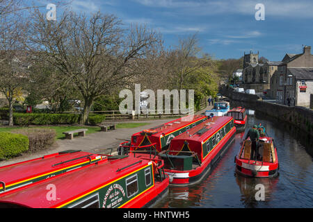 Sonnige landschaftlich schöne Aussicht auf Mann Lenkung schmalen Boot vorbei leuchtend roten Kanal Mietboote vor Anker - Leeds-Liverpool Canal, Skipton, Yorkshire, England. Stockfoto