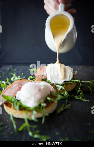 Weibliche Hand Gießen Sauce Hollandaise über Eiern Benedict Frühstück auf Schiefer Stockfoto