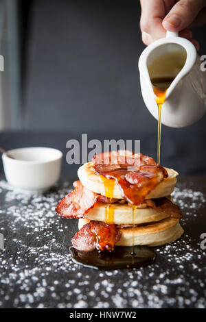 Weibliche Hand strömenden Ahornsirup über Frühstück Speck Fladen auf Schiefer Stockfoto
