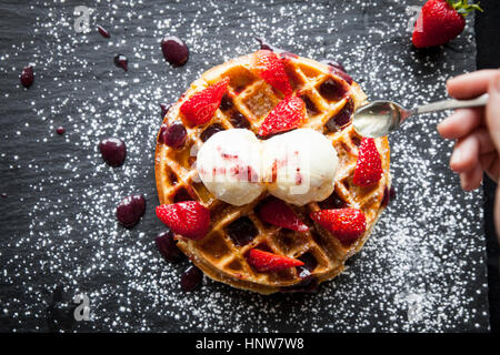 Weibliche Hand mit Löffel über Erdbeeren und Eis Waffel auf Schiefer Stockfoto