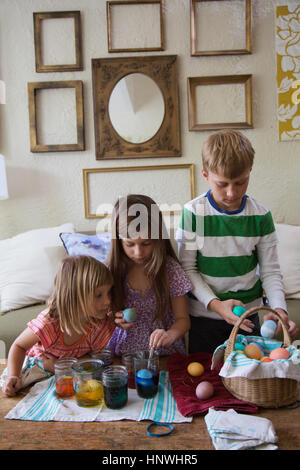 Junge und zwei Schwestern färben Ostereier in Gläsern am Tisch Stockfoto